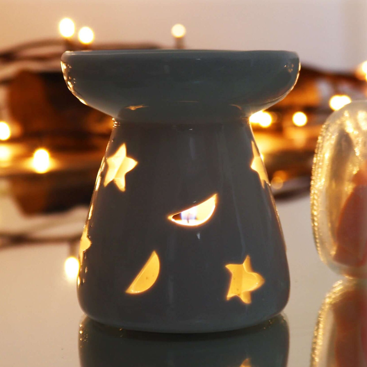 Lit ceramic oil warmer with glowing moon and star cut-outs, reflecting light in a cosy evening setting.