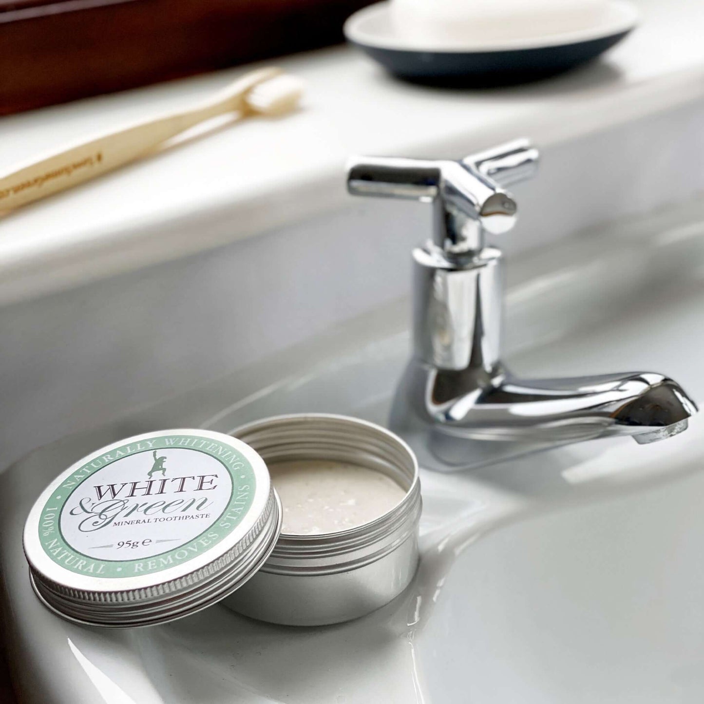 Open tin of White & Green mineral toothpaste on a bathroom sink beside a tap, with a wooden toothbrush in the background