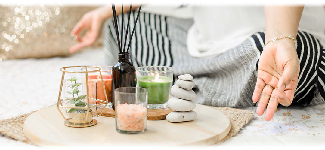 Woman meditating with candles, aromatherapy diffusers, and spa stones arranged on a wooden tray in a serene setting.