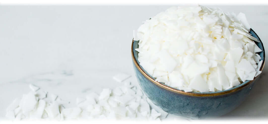 Blue ceramic bowl filled with soy wax flakes, with some flakes scattered on a white marble surface.