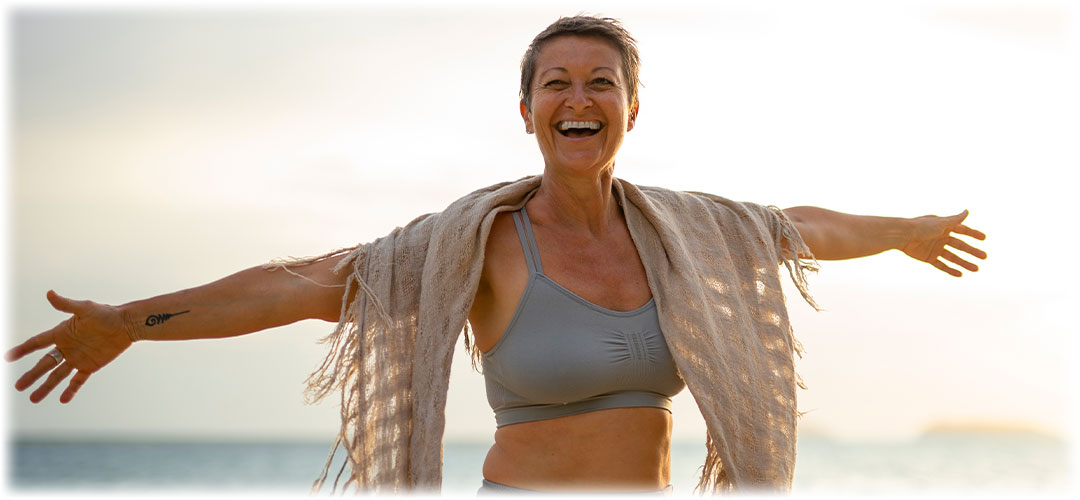 Smiling woman with short hair wearing a sports bra and shawl, standing with arms outstretched on a beach at sunset.