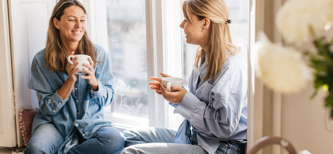 cheerful-caucasian-beautiful-women-casual-clothes-gossip-drink-tea-while-sitting-windowsill-day-leisure-lifestyle