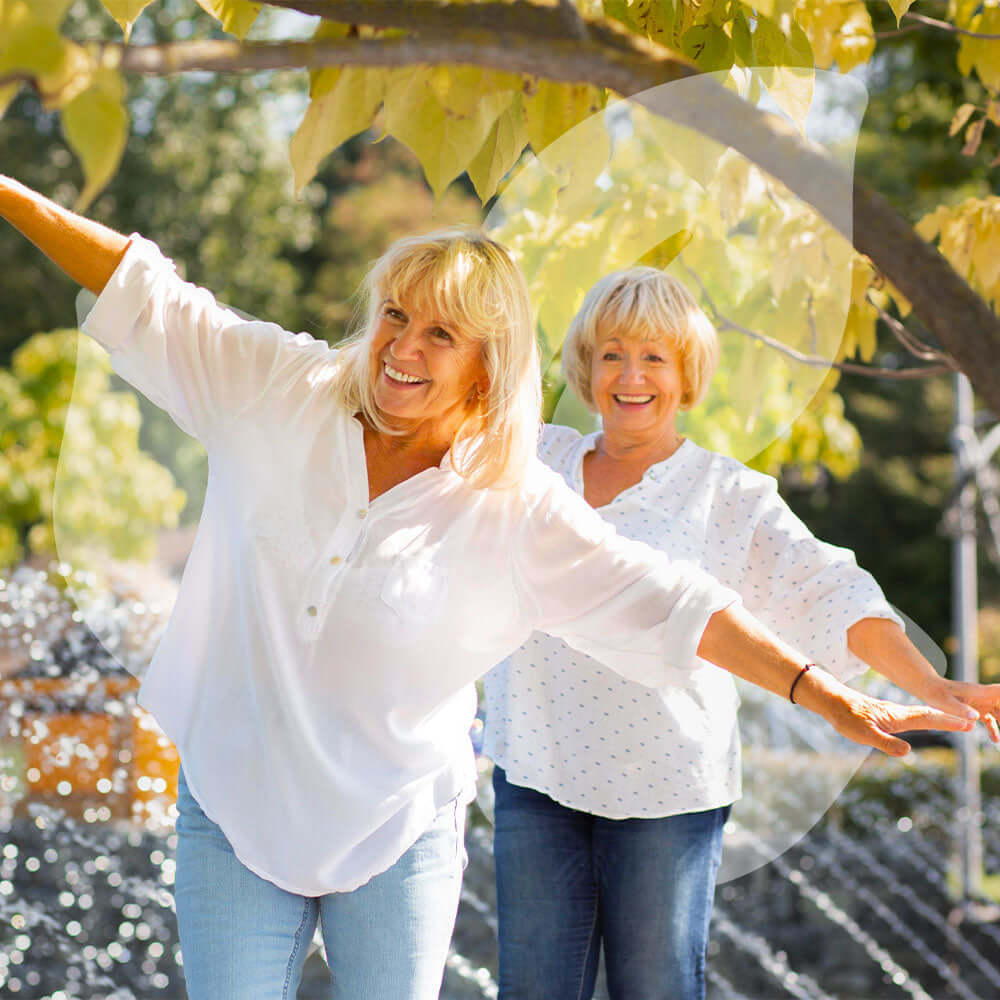 Two women in white shirts and blue jeans standing under a tree with a blurred background