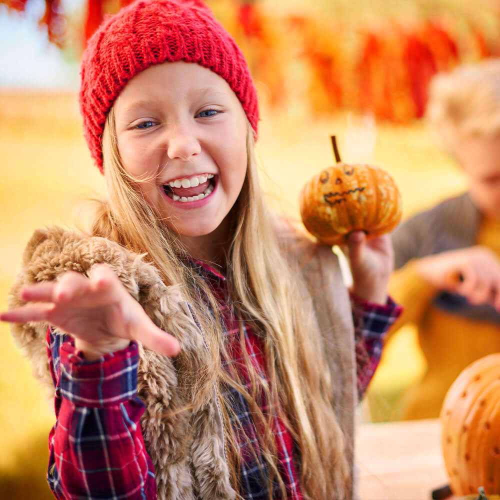 Child holding a small pumpkin with a carved face in an outdoor setting with pumpkins and autumn leaves.