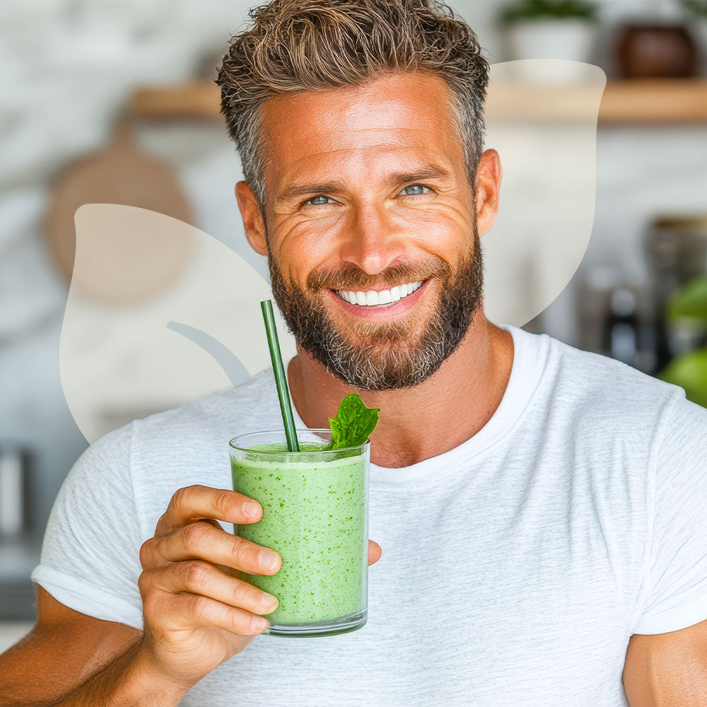 Man holding a green smoothie with a straw and mint leaf, smiling in a kitchen setting.