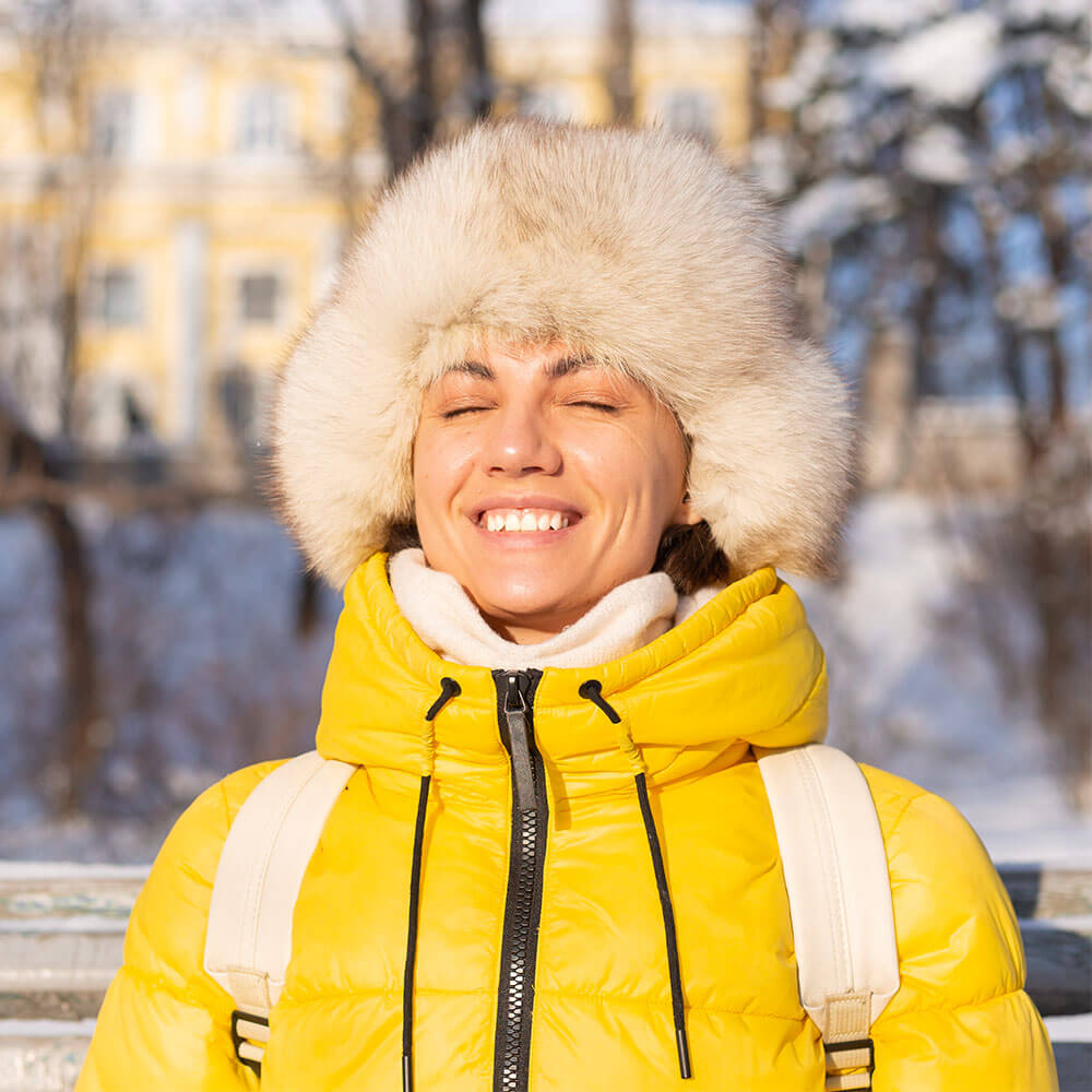 A smiling woman in a fluffy hat and yellow coat looking towards the sun on a Wintery day