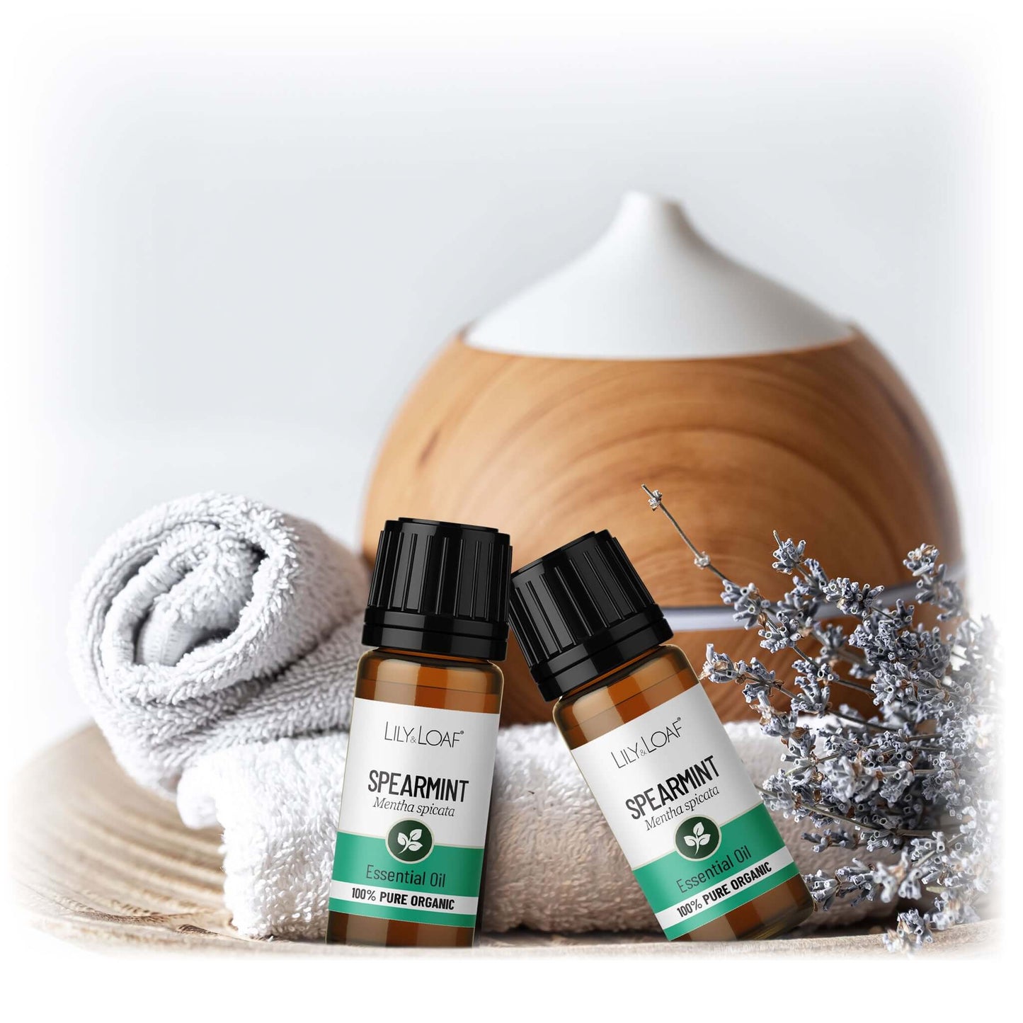 Two bottles of Lily & Loaf spearmint essential oil with a wooden diffuser and decorative elements on a light background.