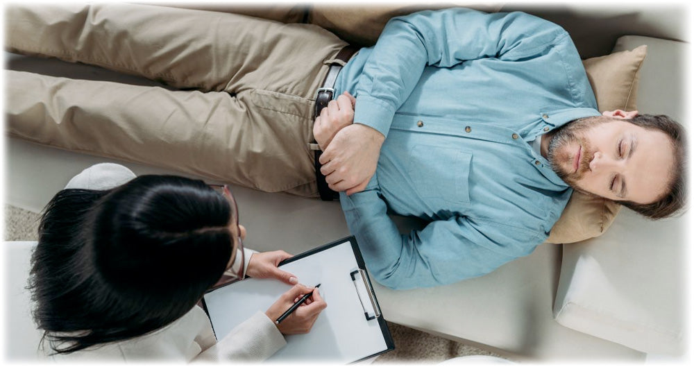 Man lying on a couch with eyes closed during a therapy session, while a therapist takes notes on a clipboard, highlighting mental health and relaxation.