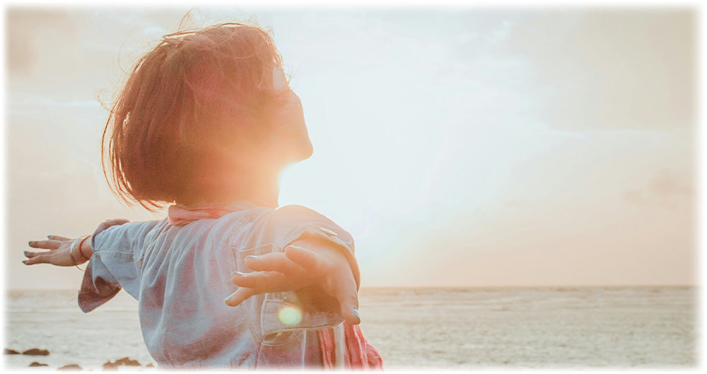 Woman standing by the ocean with arms outstretched, embracing the sunlight and fresh air, symbolizing freedom and joy.