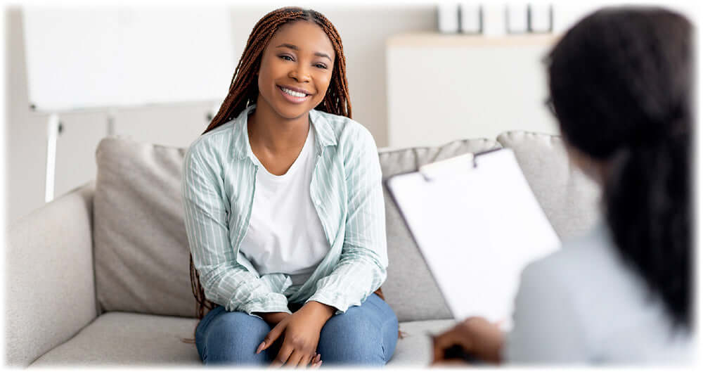 Smiling woman with braided hair sitting on a couch during a consultation with a professional holding a clipboard, representing a supportive and positive counseling session.