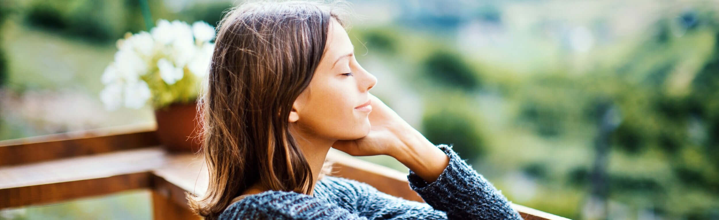 A woman with her eyes closed sitting on a wooden deck in the countryside