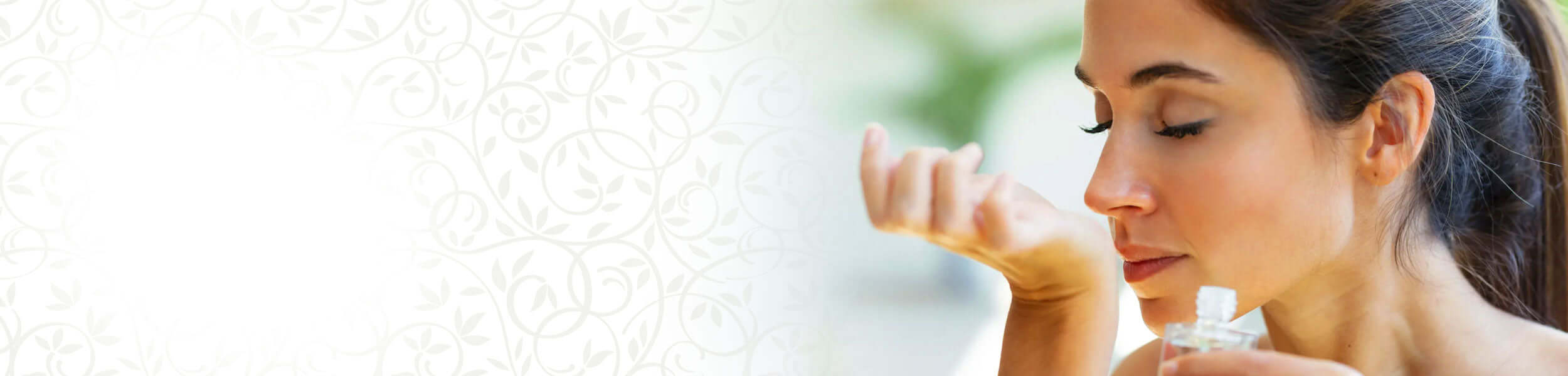Woman holding a bottle of fragrance and smelling her wrist against a soft blurred background