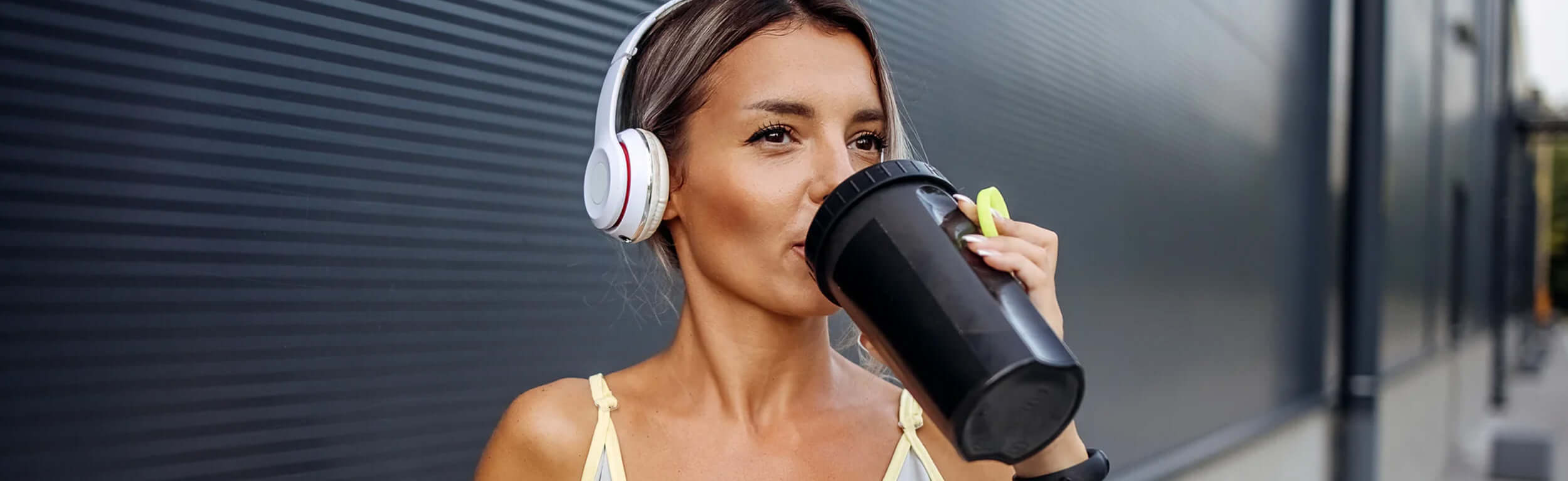 A woman wearing headphones and drinking a protein shake from a black shaker.