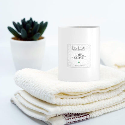 Lily & Loaf Lime & Coconut Soy Wax Candle resting on a stack of fluffy white towels with a plant in the background