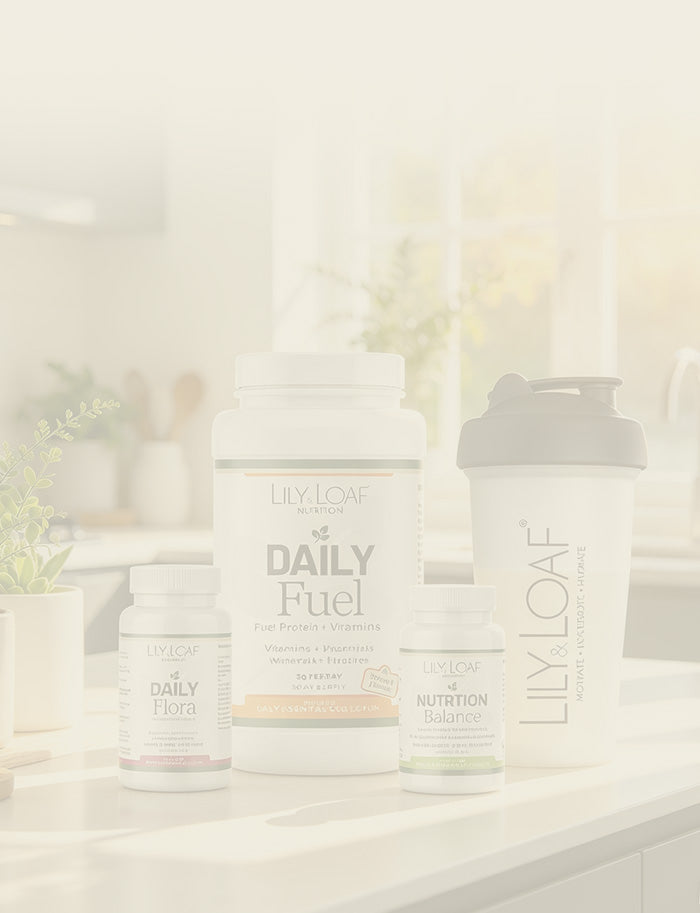 Lily & Loaf Daily Essentials supplements arranged on a kitchen counter, including a large Daily Fuel tub, a shaker bottle, a Daily Flora bottle, and a Nutrition Balance bottle, all in a bright, softly lit kitchen setting.