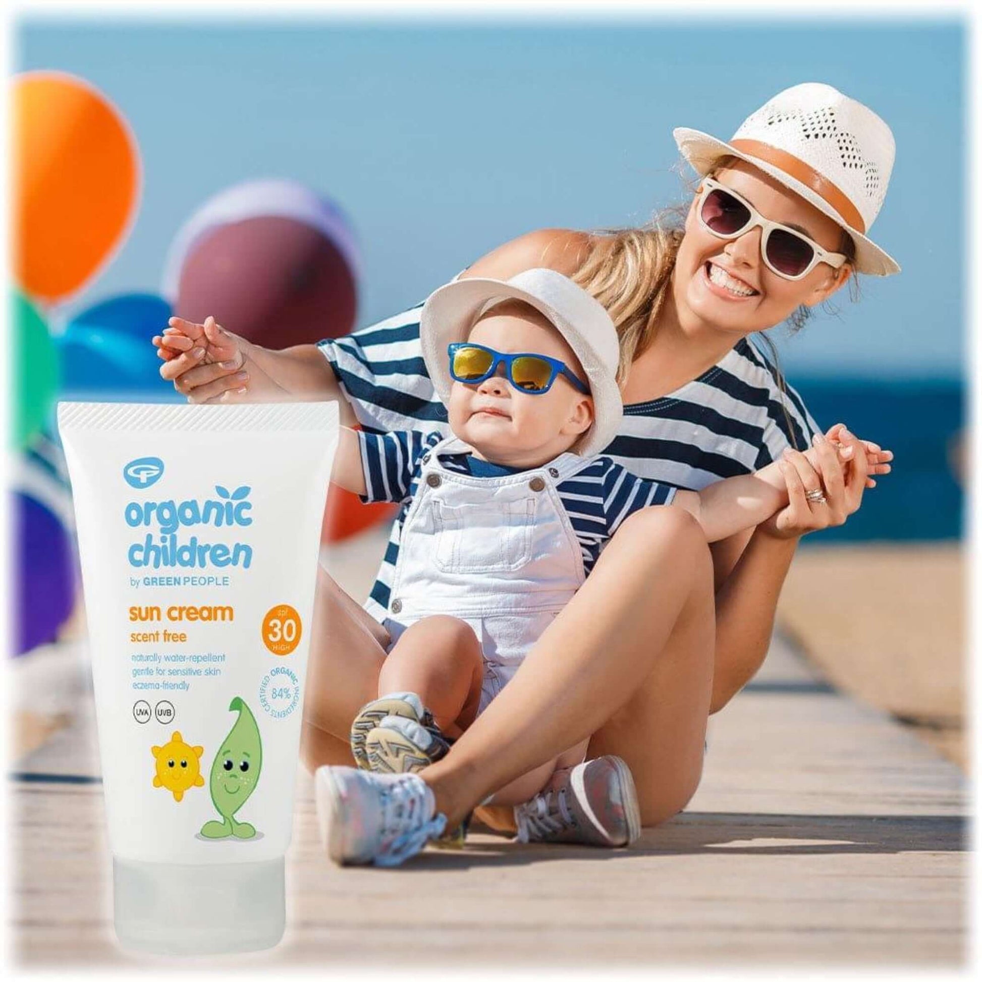 A woman and baby on a beach wearing sunglasses and hats with a bottle of Green People Organic Children’s Scent Free Sun Cream in the foreground