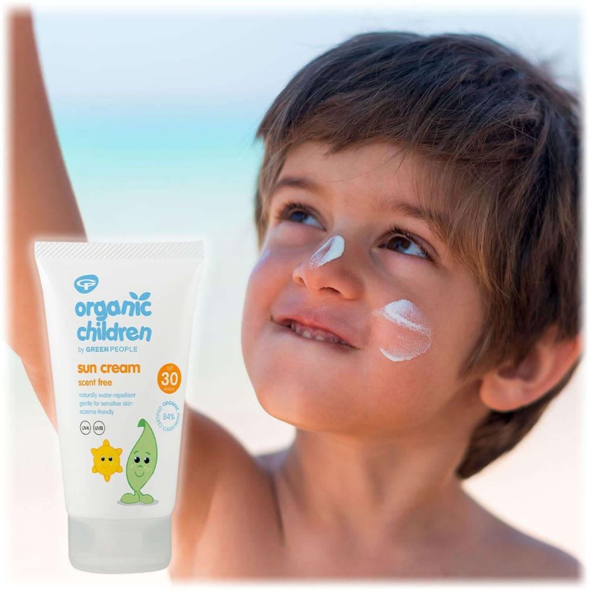 A little boy playing on the beach with sun cream on his nose and cheeks. A bottle of Green People Organic Children’s Scent Free Sun Cream SPF30 is in the foreground