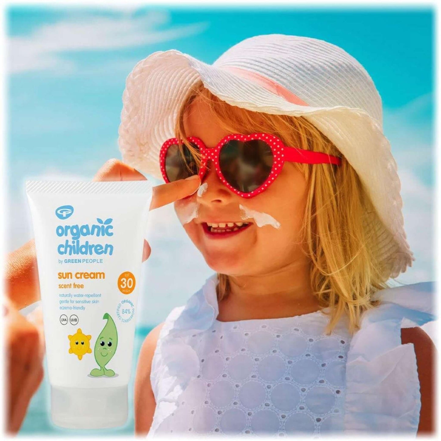 A little girl in a hat and heart sunglasses with sun cream being applied to her face, a bottle of Green People Organic Children’s Scent Free Sun Cream SPF30 is in the foreground