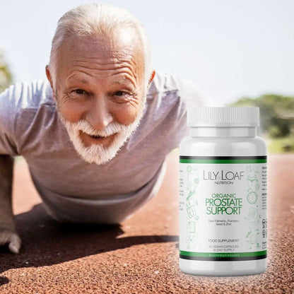 An elderly man with white hair and a white beard, smiling warmly at the camera while doing a push-up on a track. He is wearing a light grey t-shirt, and his fitness suggests an active and healthy lifestyle. Next to him is a prominently displayed bottle of Lily & Loaf's Organic Prostate Support dietary supplement