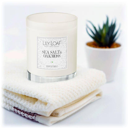 Lily & Loaf Sea Salt & Oakmoss Soy Wax Candle resting on a stack of fluffy white towels with a plant in the background