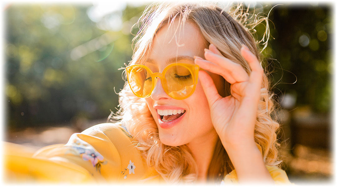 A woman with blonde curly hair wearing yellow sunglasses and a yellow top, smiling brightly and enjoying a sunny day outdoors. The background is blurred with greenery and sunlight creating a warm, cheerful ambiance.