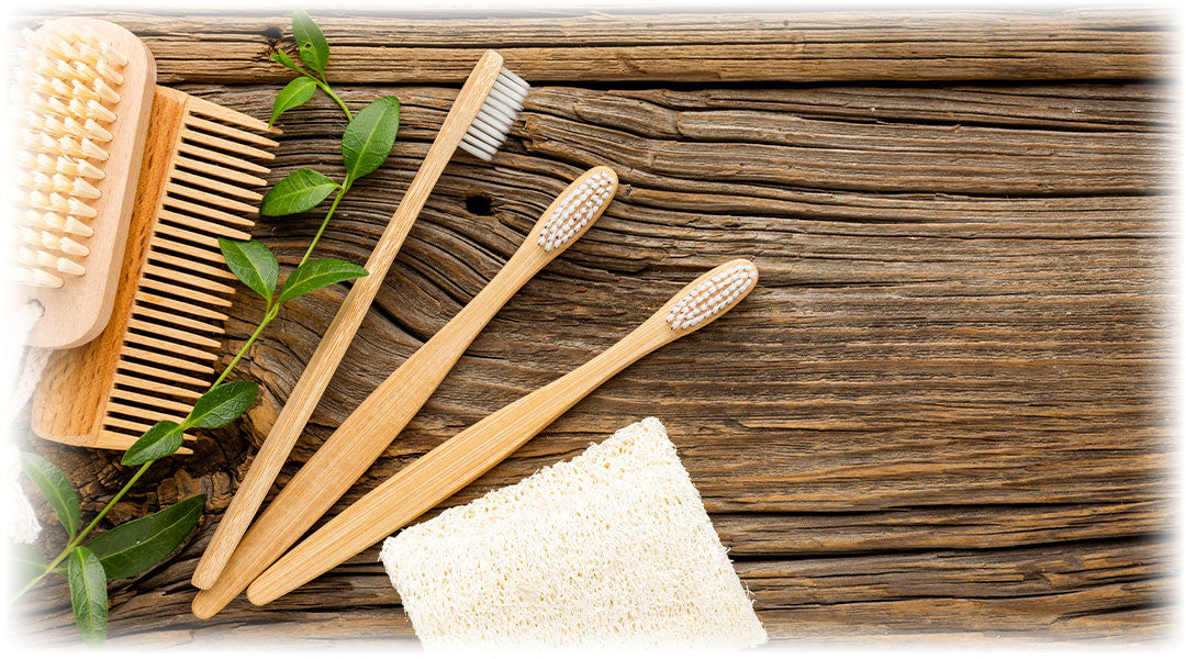 Natural self-care products including wooden toothbrushes, a comb, and a body brush, arranged on a rustic wooden surface next to green leaves and a loofah pad. Promotes eco-friendly personal hygiene.