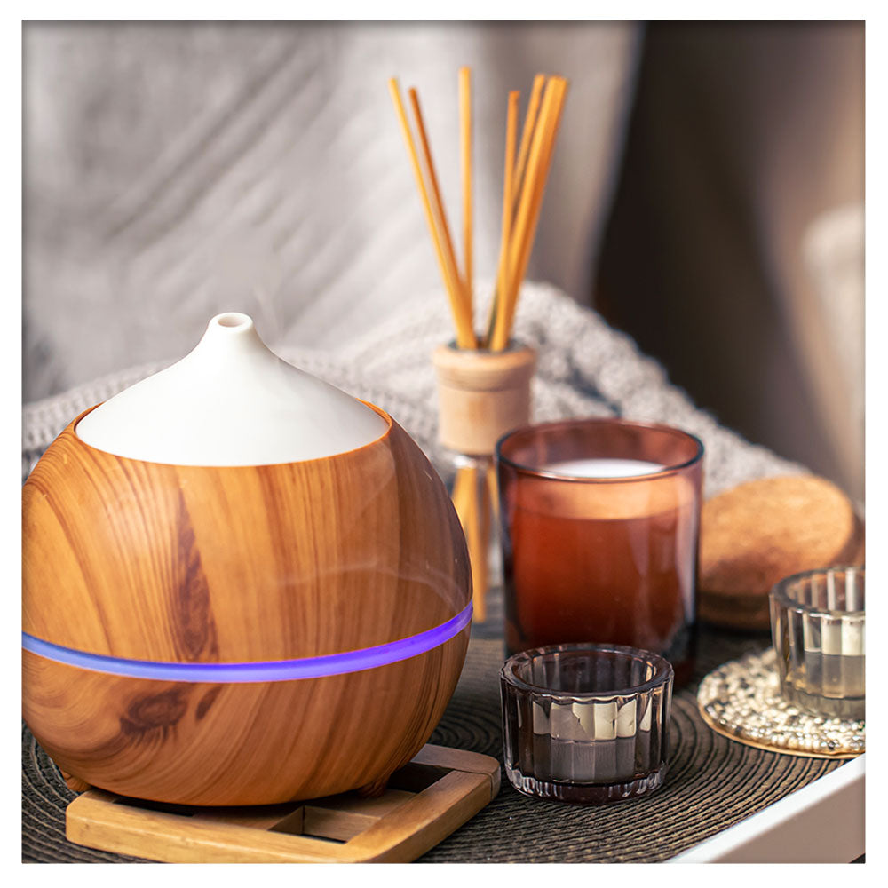 An essential oil diffuser placed on a wooden surface. The diffuser has a sleek and modern design, emitting a gentle mist. Surrounding the diffuser are candles and reed incense, adding a natural and serene touch to the scene.