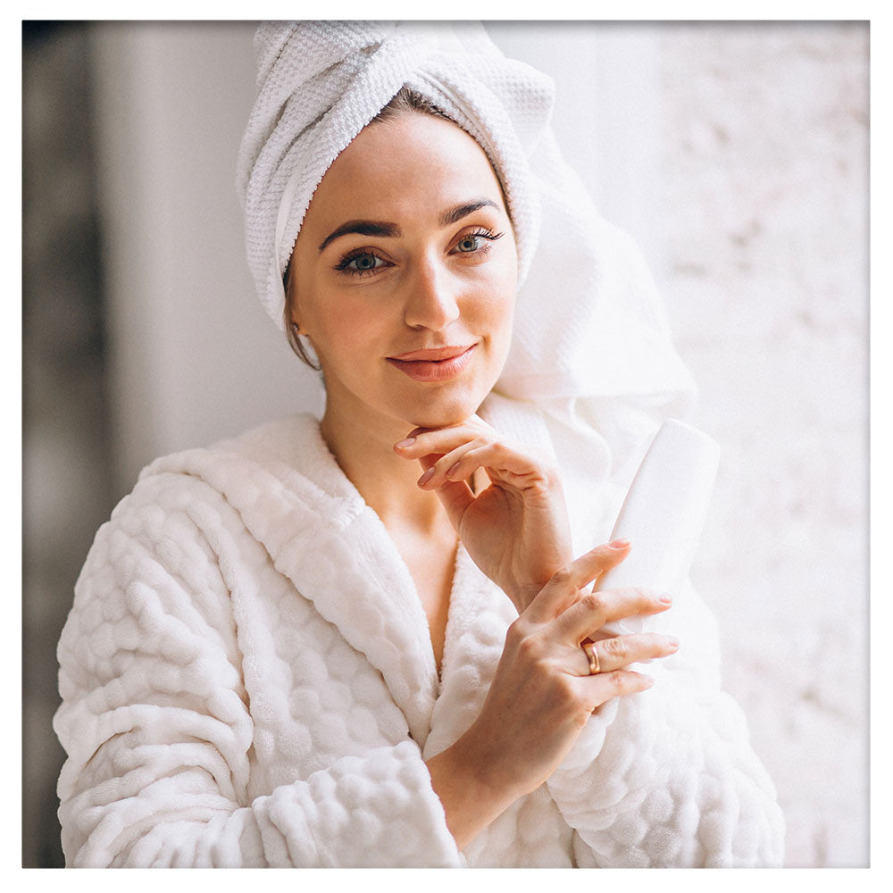 A woman with smooth, glowing skin wearing a towel robe is looking at the camera with a serene expression. The background is plain and white, emphasising her radiant complexion and natural beauty.