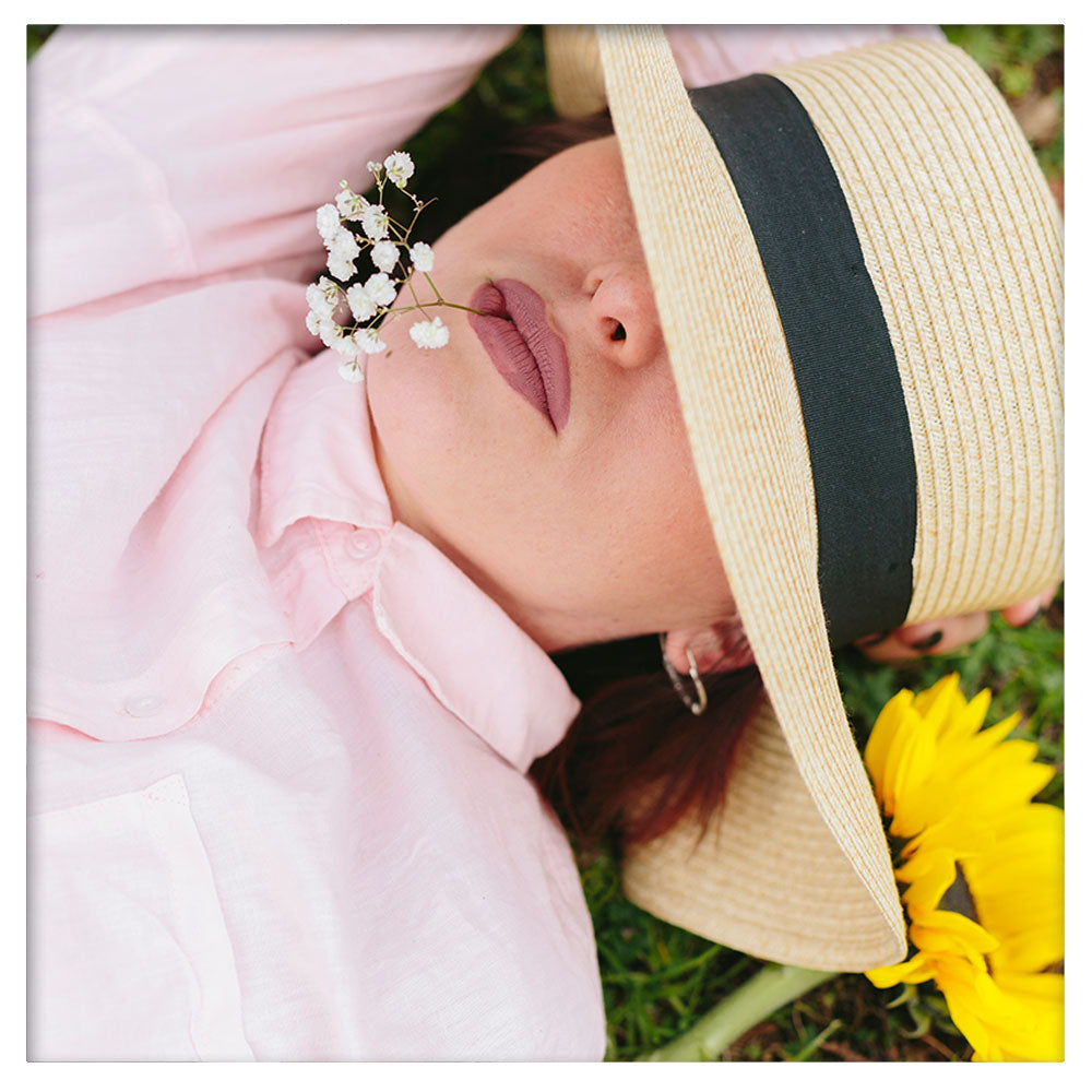A woman peacefully sleeping outdoors under a straw hat. She is lying on her back, with her face partially covered by the hat, and the background features a natural, green setting, creating a tranquil and serene atmosphere.