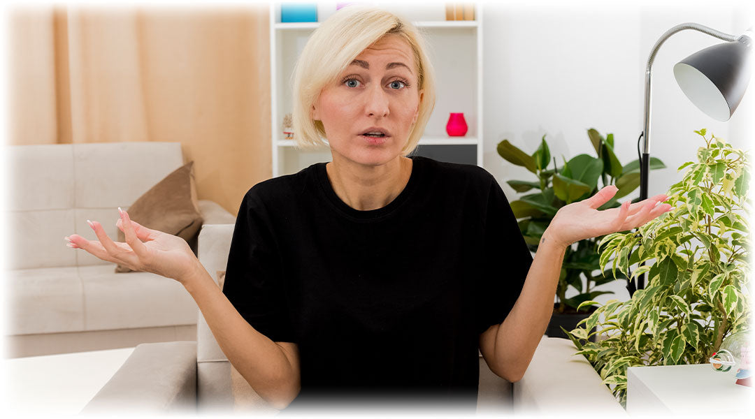 Woman with blonde hair in a black shirt shrugging her shoulders in confusion, sitting in a living room with plants and home decor.