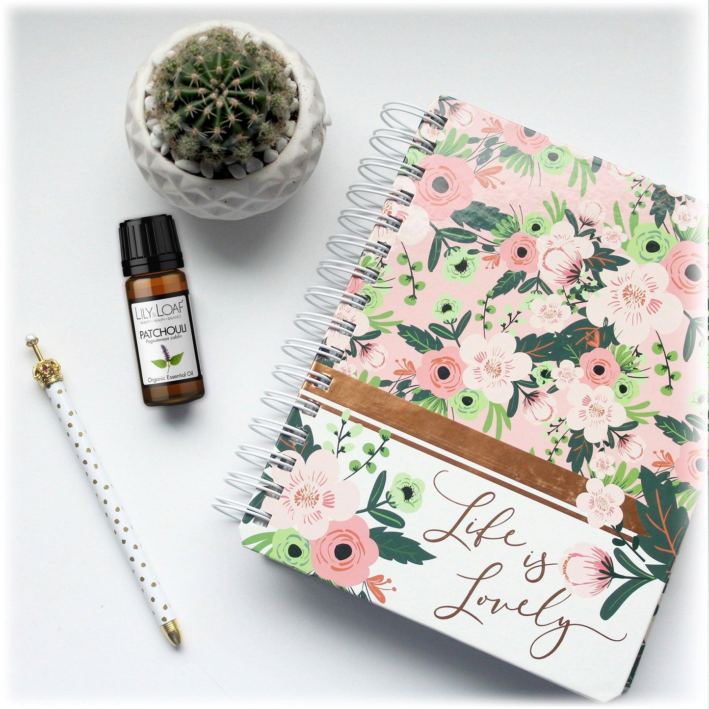 Lily & Loaf Patchouli essential oil bottle next to a floral notebook, a gold-topped pen, and a small cactus on a white surface. Promotes relaxation and creativity.