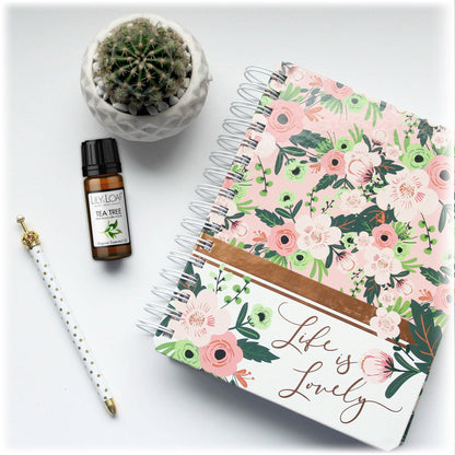 Floral notebook with 'Life is Lovely' text, Lily & Loaf Tea Tree essential oil bottle, white pen with gold polka dots, and a small potted cactus. The image promotes the soothing and refreshing benefits of Lily & Loaf Tea Tree oil for a pleasant workspace.